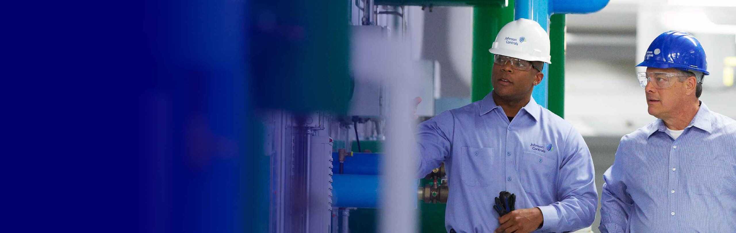 Two Johnson Controls mechanics wearing protective helmets, examining HVAC equipment in a facility